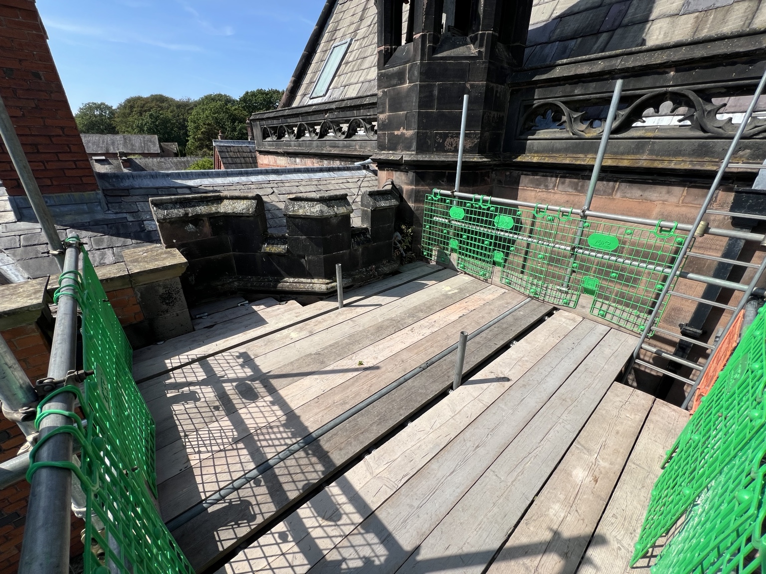 Boarded scaffold platform with green edge panels beside stone parapets and roof details at Arley Hall